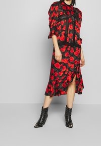 Schwarzes Blumenkleid mit roten Blumen, gerafftem Saum und einem seitlichen Schlitz. Kombiniert mit schwarzen, genoppten Ankle Boots. Strukturierter Stoff, strukturiertes Design.