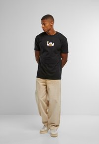 Svart t-skjorte med en tegneserieillustrasjon av en katt med en stabel pannekaker. Modell iført lysebeige bukser og lyse sneakers.
