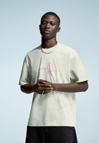Young man in light beige T-shirt with pink star graphic, black pants, and silver chain necklace, standing against a light blue background.