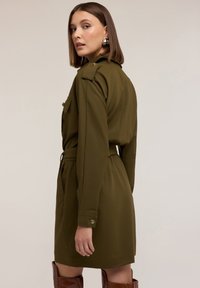 Abito camicia verde oliva con maniche lunghe, polsini bottonati e vita con cintura. Presenta due tasche sul petto e una texture liscia e morbida.