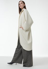 Abrigo largo beige con una textura suave, mangas anchas y bolsillos, usado sobre pantalones de pierna ancha oscuros y estampados y tacones negros puntiagudos.