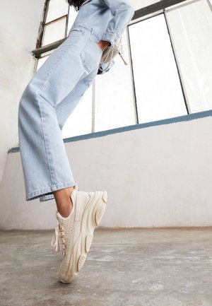 Personne portant un jean en denim bleu clair et des baskets beige épaisses, se tenant sur la pointe des pieds dans une pièce lumineuse de style industriel.