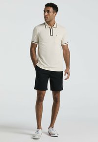 Polo beige avec fermeture zippée, col noir et bord de manches assorti, associé à un short noir et des baskets blanches avec des accents rouges.