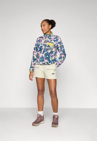 Fleece-Pullover mit buntem Blumenmuster, gelbe Brusttasche; beige Shorts mit Seitentaschen; braune Wanderschuhe mit weißen Socken.