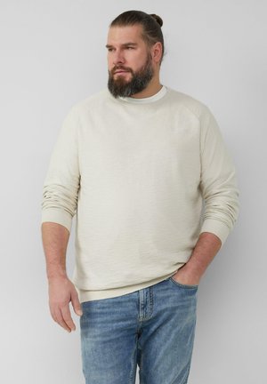 Helle beige Strickjacke mit strukturiertem Stoff, Rundhalsausschnitt und gerippten Bündchen. Kombiniert mit blauen Jeans.