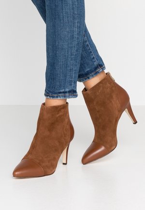 Botas de ante marrón hasta el tobillo con puntas de cuero afiladas y tacones de 6 cm. Detalle de cremallera lateral y textura suave. Combinadas con jeans azules con dobladillo.