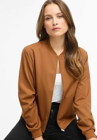 Jachetă bomber maro cu închidere cu fermoar, guler striat și mâneci lungi; purtată peste un top alb, asortată cu blugi negri.