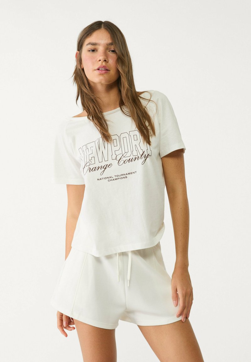 T-shirt blanc en coton avec col rond et manches courtes, arborant l'inscription "NEWPORT Orange County" en imprimé marron. Associé à un short blanc à cordon de serrage.