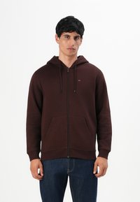 Hombre vestido con una sudadera con cremallera de color marrón oscuro, con bolsillos frontales y vaqueros azules, de pie contra un fondo blanco liso.