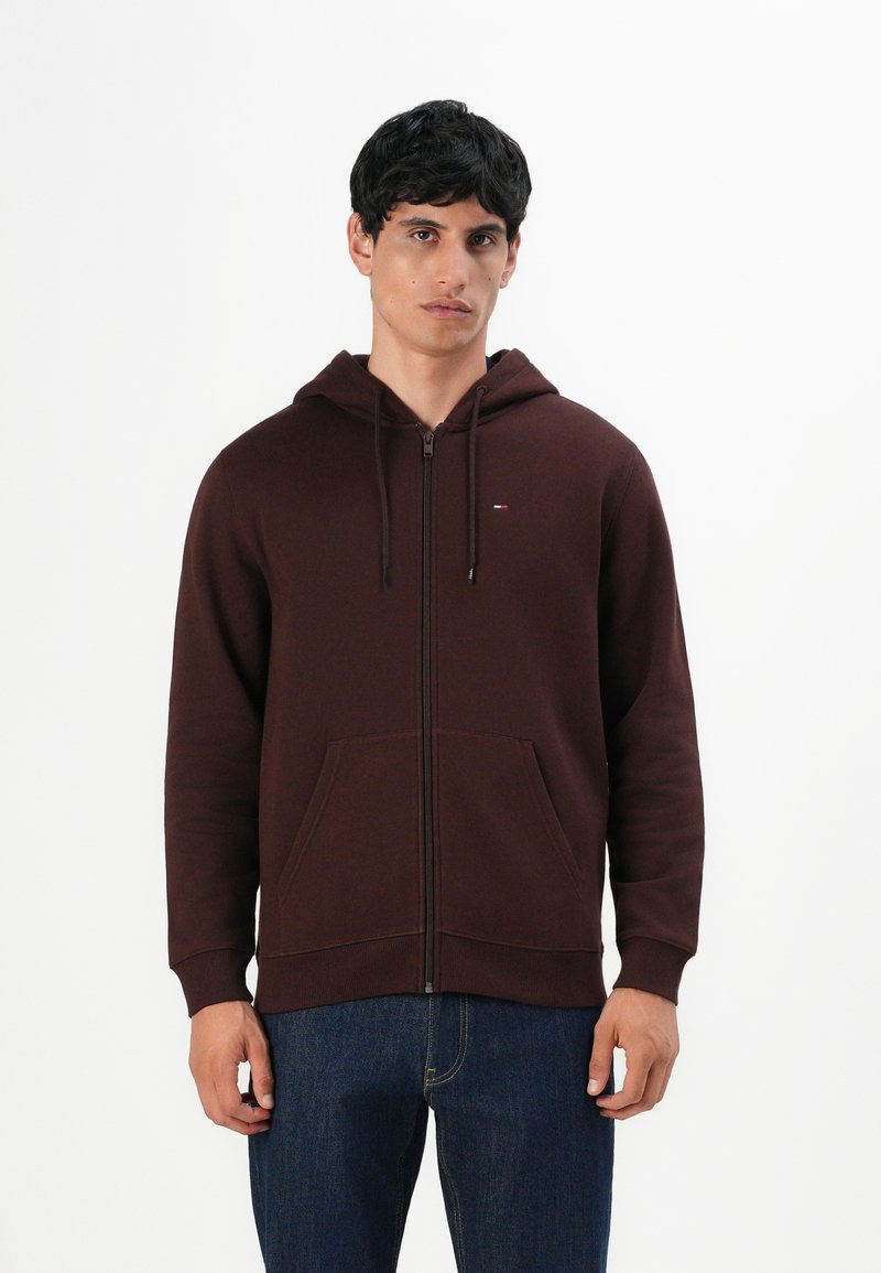 Hombre vestido con una sudadera con cremallera de color marrón oscuro, con bolsillos frontales y vaqueros azules, de pie contra un fondo blanco liso.