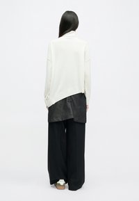 Κρέμα oversized πουλόβερ με διπλωμένο γιακά, φορεμένο πάνω από μια μαύρη τουνίκ, συνδυασμένο με φαρδύ μαύρο παντελόνι και μπεζ αθλητικά παπούτσια.