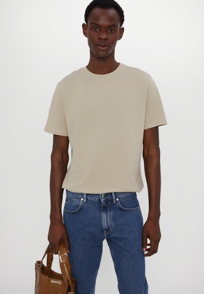 Camiseta de algodón beige con cuello redondo, mangas cortas, combinada con jeans de mezclilla azules y un bolso tote brillante de color marrón.