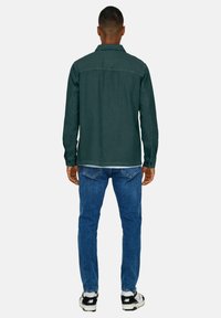 Dunkelgrünhe Hemd mit Kragen, langen Ärmeln und geradem Schnitt, kombiniert mit blauen Jeans und schwarzen Sneakern.