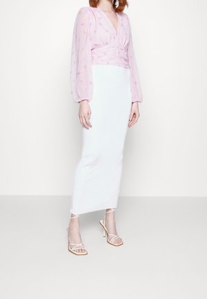 Vrouw die een lange witte rok draagt, een doorschijnende roze bloemenblouse met lange mouwen en witte sandaaltjes met hoge hak en bandjes, staand tegen een witte muur.