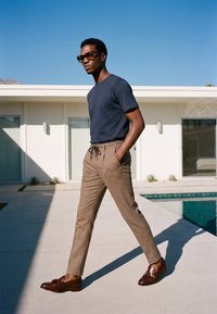 Homme portant des lunettes de soleil, un t-shirt bleu marine, un pantalon marron à carreaux et des mocassins marron, marchant à côté d'un bâtiment blanc moderne avec une piscine sous un ciel clair.
