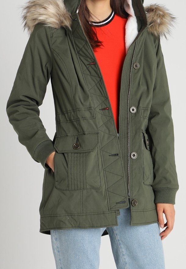 Kvinna iklädd en grön parkas med pälsluva, röd tröja och ljusblå jeans, står med händerna i fickorna mot en enfärgad bakgrund.