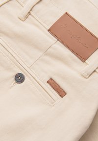 Gros plan sur la poche d’un pantalon en denim beige avec un bouton en métal et deux étiquettes de marque en cuir marron cousues près de la poche.