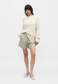 Γυναίκα που φοράει κρεμ πλεκτό πουλόβερ, ανοιχτό γκρι σορτς με κορδόνι και μπεζ μυτερές μπαλαρίνες σε απλό λευκό φόντο.