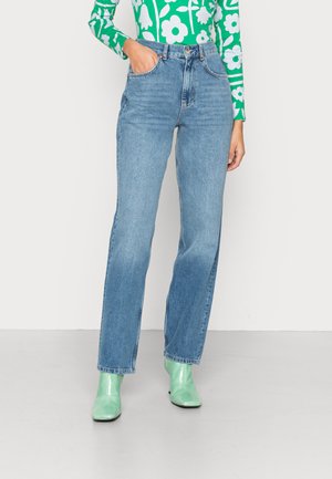 Jeans de ganga azuis de cintura alta com corte reto, apresentando cinco bolsos e desbotamento subtil. Combinados com botins verdes.
