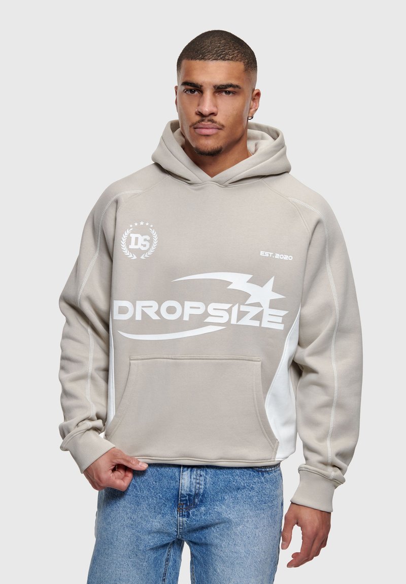 Homme portant un sweat à capuche beige avec un logo "Dropsize" blanc et un jean bleu clair, debout devant un fond gris clair uni.