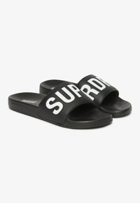 Sorte slidesandaler lavet af gummi med en bred rem, der har ordet "SUPREME" i fede hvide bogstaver. Tekstureret sål for bedre greb.