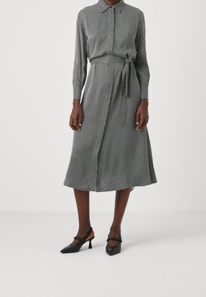 Donna che indossa un vestito midi a maniche lunghe, con motivo bianco e nero, cintura annodata in vita e scarpe slingback nere con tacco.