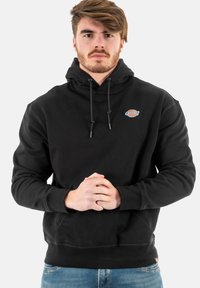 Schwarzer Kapuzenpullover mit einer frontalen Tasche, Kordeln und einem kleinen Logo. Der Stoff wirkt weich, lässige Passform mit gerippten Bündchen und Saum.