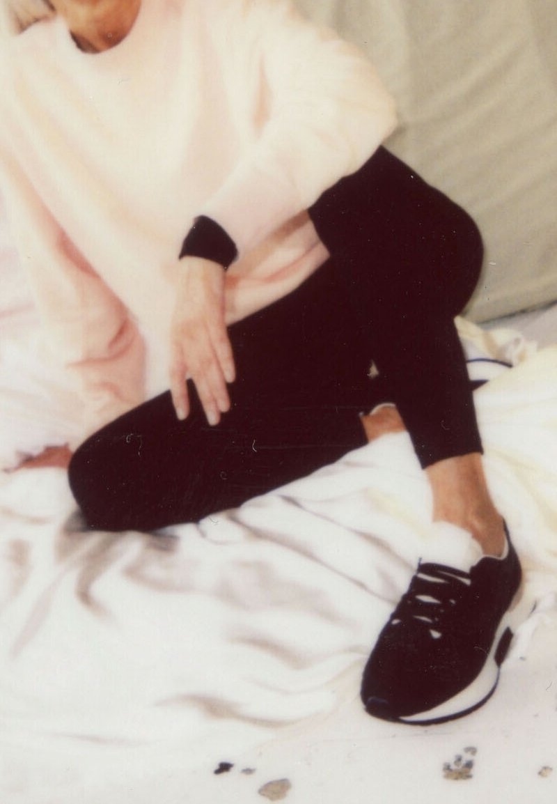Personne assise en tailleur sur un lit blanc, portant un pull rose clair, des leggings noirs et des baskets noires avec des semelles blanches.
