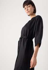 Μαύρο λινό φόρεμα με τρία τέταρτα μανίκια και ζώνη δέσιμο στη μέση. Διαθέτει απλή λαιμόκοψη και άνετη γραμμή, αναδεικνύοντας μια υφή υφάσματος.