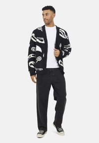 Zwart-wit geblokt cardigan van fluffy materiaal, met abstracte ontwerpen, gedragen over een wit T-shirt en zwarte broek.