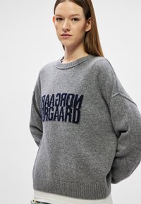 Suéter cinza de malha com um decote redondo, ombros caídos e um padrão de logótipo azul-marinho na frente. Mangas e bainha com textura canelada.