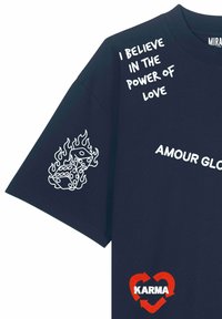 Maglietta blu navy con testo bianco "Credo nel potere dell'amore", grafica di dadi infuocati sulla manica e frecce a forma di cuore rosso con "Karma" sulla parte frontale inferiore.