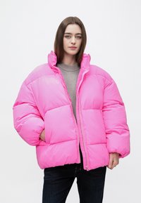 Žena v jasne ružovej veľkej prešívanej bunde cez sivý sveter a tmavé džínsy, stojaca pred jednoduchým bielym pozadím.