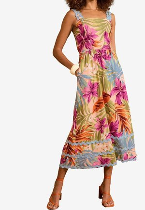 Kvinde iført en ærmeløs midikjole med blomsterprint med bølgede kanter og orange højhælede sandaler med stropper.