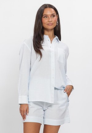 Vrouw met lang donker haar die een lichtblauw gestructureerd overhemd met knopen en bijpassende korte broek draagt, staand met één hand in de zak.
