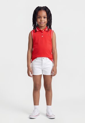 Mladé dievča s zatočenými vlasmi, oblečená v červenej bezrukávovej polokošeli, bielych šortkách, bielych ponožkách a bielych teniskách, stojaca čelom vpred.