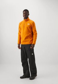 Oranžen pulover iz flisa z ovratnikom na polovični zadrgi, kombiniran s črnimi nepremočljivimi hlačami z žepi in logotipi.