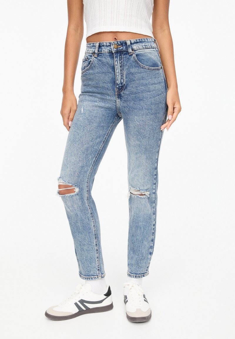 Femme portant un jean bleu délavé taille haute avec genoux déchirés, un top blanc court sans manches, des chaussettes blanches et des baskets blanches avec des rayures noires.