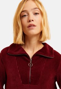 Vestito in corduroy bordeaux con zip frontale e colletto oversized, caratterizzato da un motivo a coste testurizzato e una zip circolare.