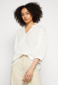 Blusa leve e branca com decote em V, textura de bolinhas e mangas curtas volumosas. Combinada com calças bege de cintura alta.