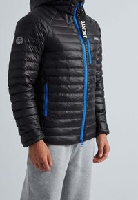 Veste matelassée noire avec capuche, coutures verticales, fermeture éclair bleue et détail logo sur la poitrine et la manche. Texture lisse et brillante.