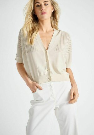 Femme blonde portant un cardigan beige clair boutonné avec des détails perforés et un pantalon blanc large, posant avec une main dans la poche.