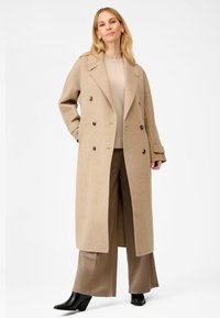 Beige dobbeltbrystet uldfrakke med bred krave, forlommer og brune knapper, båret over en beige top og brune bukser. Sorte hælede ankelstøvler.