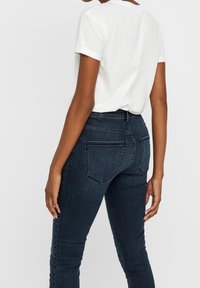 Vaqueros de mezclilla en azul oscuro, de alta cintura y con una silueta ajustada. Cuentan con bolsillos traseros y una textura suave, combinados con una camiseta blanca.