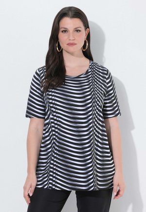 Frau mit langen dunklen Haaren, die große Creolen trägt und ein schwarz-weiß gestreiftes Kurzarmshirt, stehend vor einer weißen Wand.