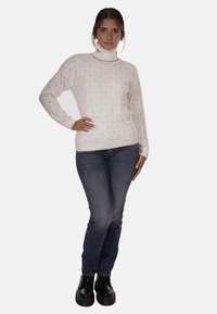 Maglione di colore crema con un motivo a trama, collo alto e polsini a costina, abbinato a jeans blu scuro e stivali neri con lacci.