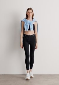 Čierny crop top a legíny značky Calvin Klein v kombinácii so svetlomodrým sveterom previazaným okolo ramien, doplnené bielymi teniskami.