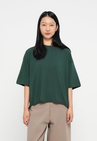 Fiatal nő egyenes fekete hajjal, laza sötétzöld rövid ujjú felsőt és világos bézs nadrágot visel egy sima fehér háttér előtt.