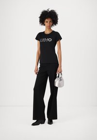 Black short-sleeve t-shirt with "LIU JO" in metallic lettering, paired with wide-leg black trousers and a small silver handbag.