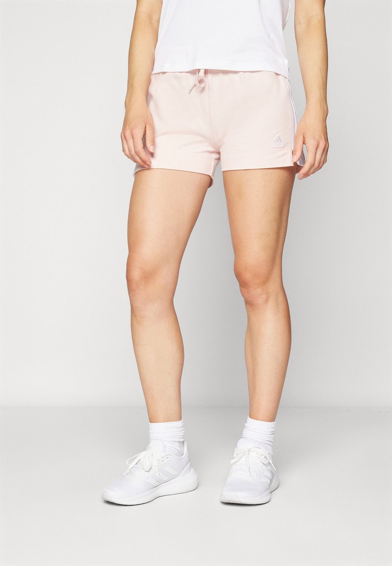 Femme portant un short de sport rose pâle, des chaussettes blanches et des baskets blanches, debout devant un fond clair uni.